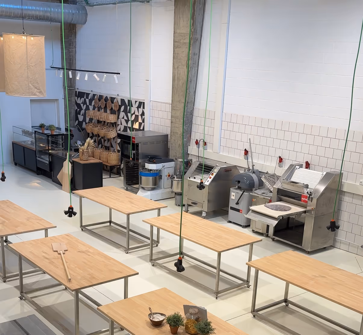 Interior de cocina industrial con mesas de trabajo de madera, maquinaria para panadería y estantes con cestas decorativas.