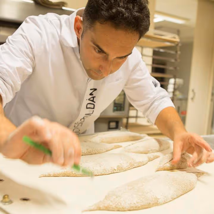 Panadero con uniforme blanco marcando masa de pan en una mesa de trabajo en panadería.