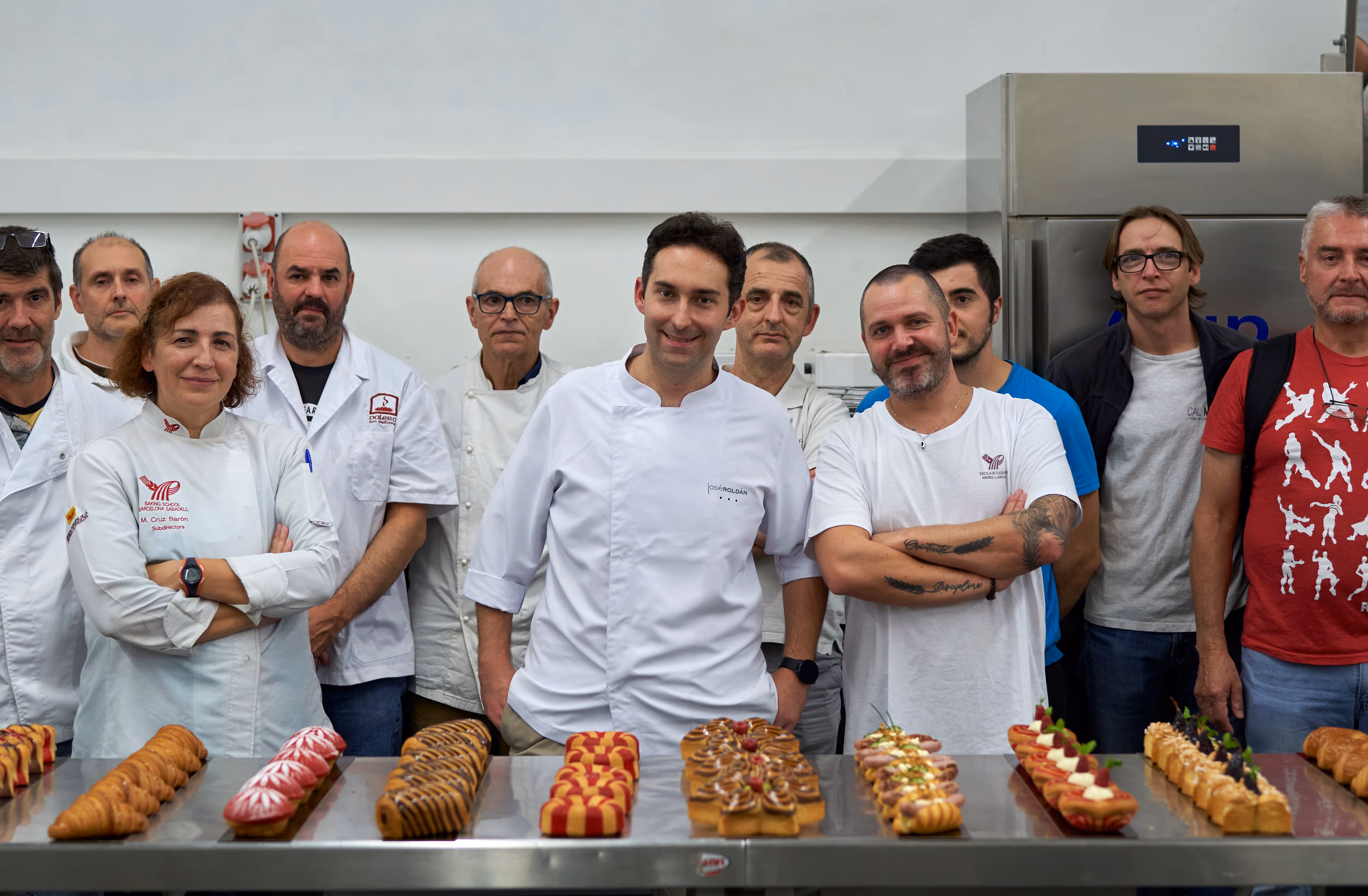 Un grupo de pasteleros y chefs posan detrás de una mesa con una variedad de pasteles elaborados y decorados en una cocina profesional.