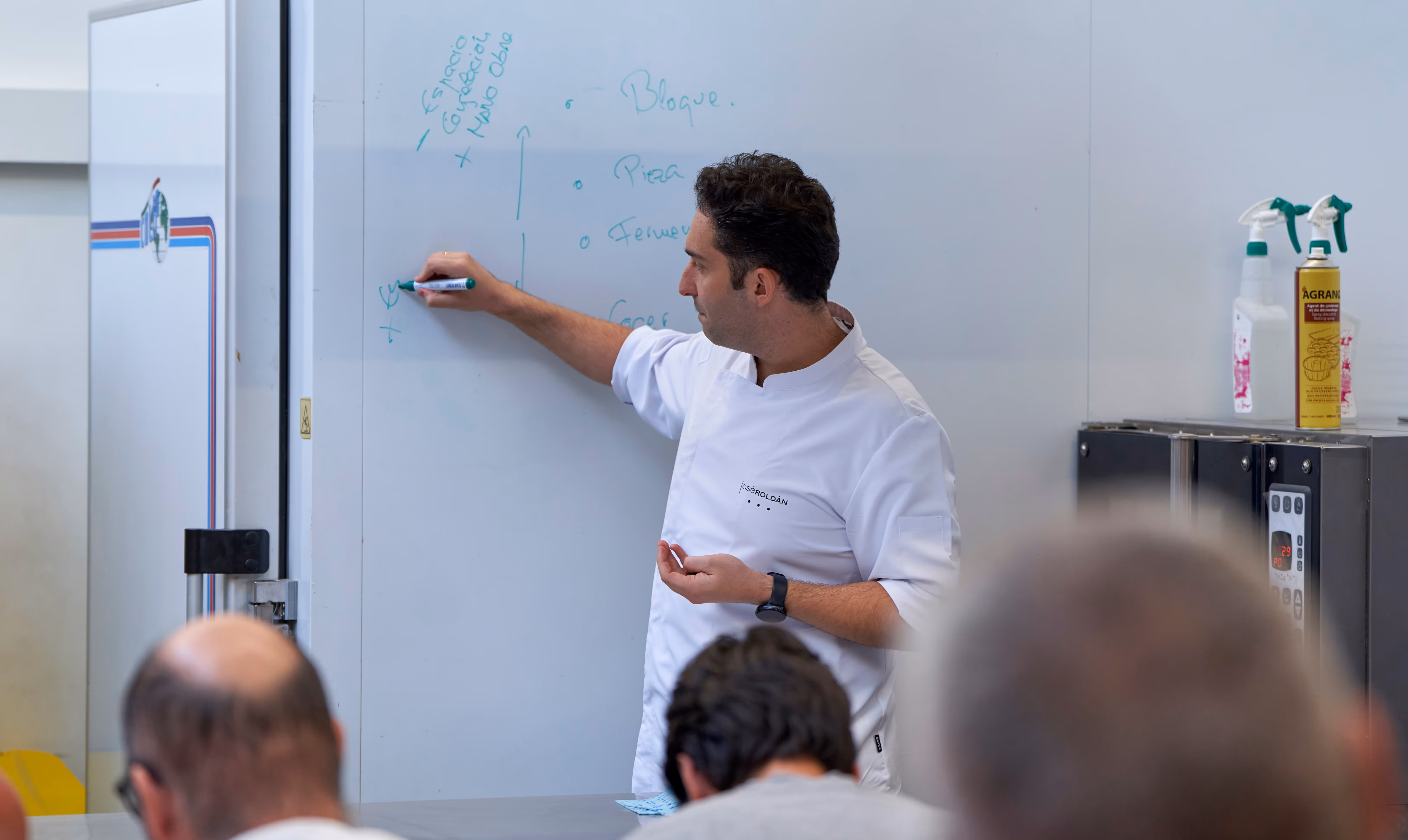 Chef with white uniform writing notes on a whiteboard during a class with attendees in foreground.