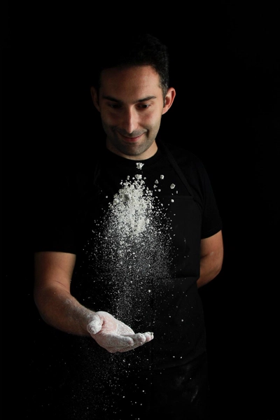 Hombre con camiseta y delantal negros lanzando harina al aire con una mano abierta.