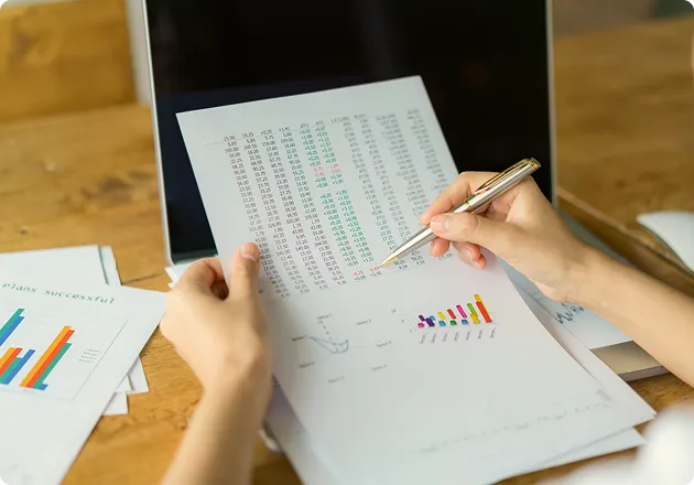 Person holding a pen and reviewing printed financial charts and data in front of a laptop on a wooden desk.