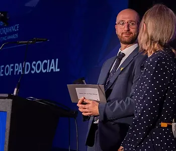 Man in a suit holding an envelope and woman in a polka dot dress standing on stage during an event.
