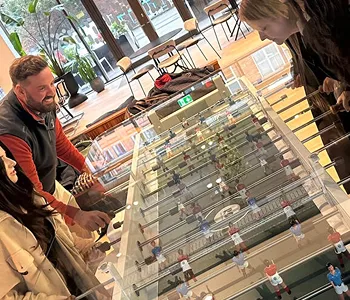 Group of four people enjoying a foosball game around a table in a bright room with large windows.