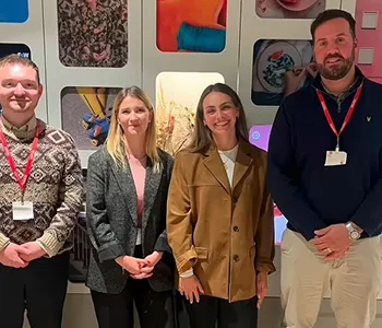 Four people standing in a row in front of a wall with colorful framed artwork, smiling at the camera.