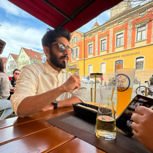 Man wearing sunglasses and a light shirt sitting at a wooden table outdoors, holding a glass of white wine, with colorful buildings in the background.