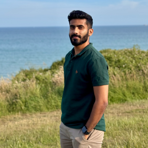 Man with beard wearing a dark green polo shirt standing on grass with the ocean and blue sky in the background.