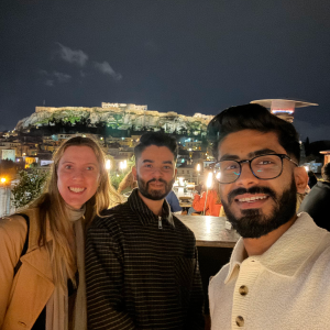 Three friends smiling for a selfie at night with the illuminated Acropolis in Athens in the background.