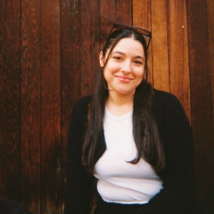 Smiling young woman with long dark hair, wearing glasses on her head, white shirt, and black cardigan, standing against wooden wall.