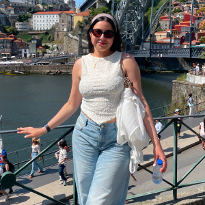 Woman in sunglasses and white sleeveless top posing by a riverside railing with a large metal bridge and colorful buildings in the background.