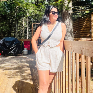 Woman wearing white sleeveless top and shorts standing on wooden deck next to railing with trees in the background.