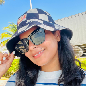 Smiling woman wearing a black and white checkered bucket hat and large black sunglasses outdoors with a clear blue sky.