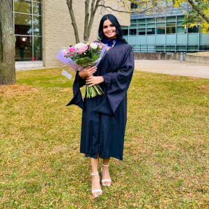 Smiling woman in a black graduation gown holding a bouquet of pink flowers standing on grass outside a building.