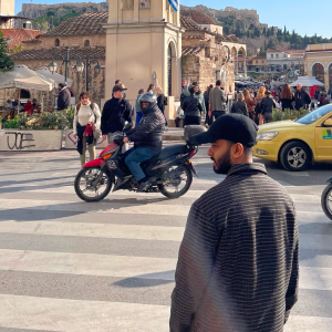 Busy city street scene with people walking, a motorcyclist driving, and a man in a black cap in the foreground.