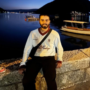 Man with a beard wearing a white sweatshirt and black pants, sitting on a stone ledge near a calm water body with boats at night.
