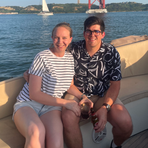 Smiling couple sitting on a boat with water, a sailboat, and a red bridge in the background.