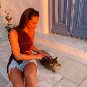Woman in a burgundy top and denim shorts sitting on stone steps petting a brown tabby cat at sunset.