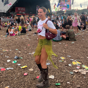 Woman smiling and posing in a festival crowd wearing a white cropped t-shirt, green fringe skirt, and brown boots.