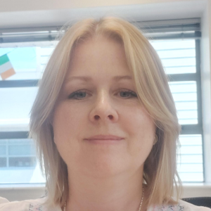 Smiling woman with shoulder-length blonde hair in front of a window with a blurred Irish flag.