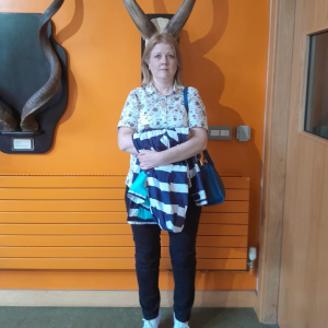 Woman standing indoors in front of an orange wall with deer antlers mounted behind her, giving the illusion that the antlers are on her head.