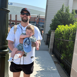 Man wearing sunglasses and a baseball cap holding a toddler outside near a building with greenery and a streetlamp.