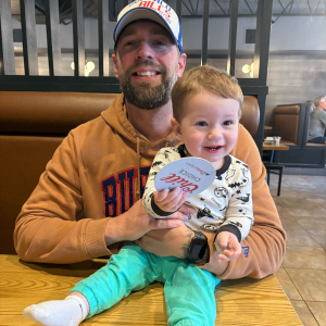 Man wearing a cap and orange hoodie holding a smiling baby dressed in a white patterned shirt and teal pants at a restaurant table.