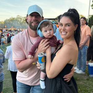 Smiling man and woman holding a baby wearing blue headphones at an outdoor event with a crowd in the background.