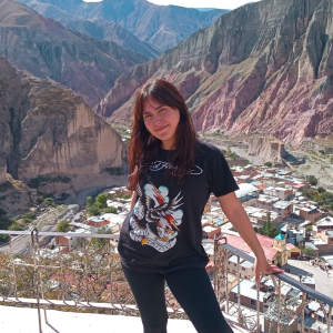 Young woman with long brown hair wearing a black graphic t-shirt, standing outdoors with a mountainous village landscape in the background.