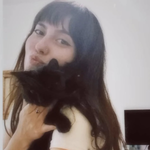 Young woman with long dark hair holding a black cat close to her face and smiling softly.