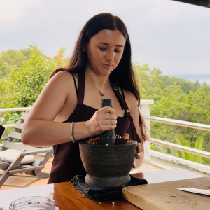 Woman using a mortar and pestle to grind ingredients at an outdoor table with greenery in the background.