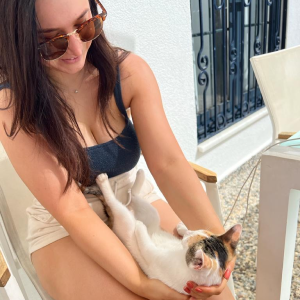 Woman wearing sunglasses and a dark tank top sitting on a chair, gently holding and looking at a small calico cat in her lap.