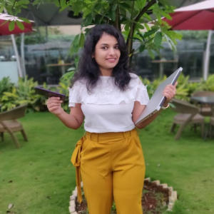 Young woman in white blouse and mustard pants holding a tablet and notebook, standing outdoors in a garden area.