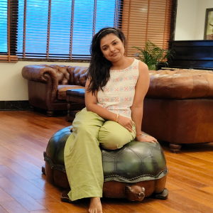 Smiling woman with long dark hair sitting barefoot on a green leather ottoman in a cozy living room with wooden floors and brown leather sofas.