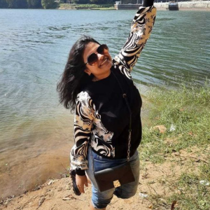 Smiling woman in sunglasses and patterned jacket raising one arm beside a calm lake shore.