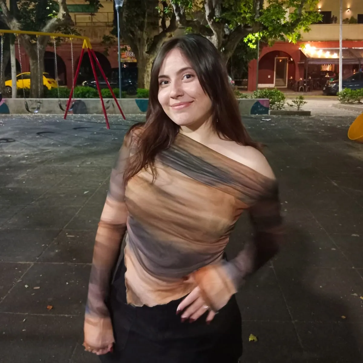 Young woman with long brown hair smiling, wearing a sheer, off-shoulder top in brown and black tones, standing in a nighttime playground.