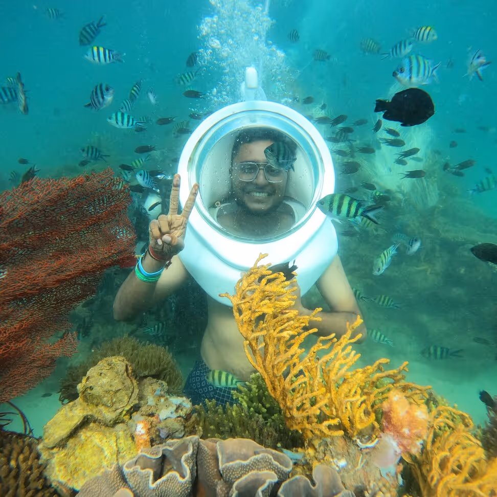 Smiling man wearing an underwater helmet making a peace sign surrounded by colorful coral and tropical fish.