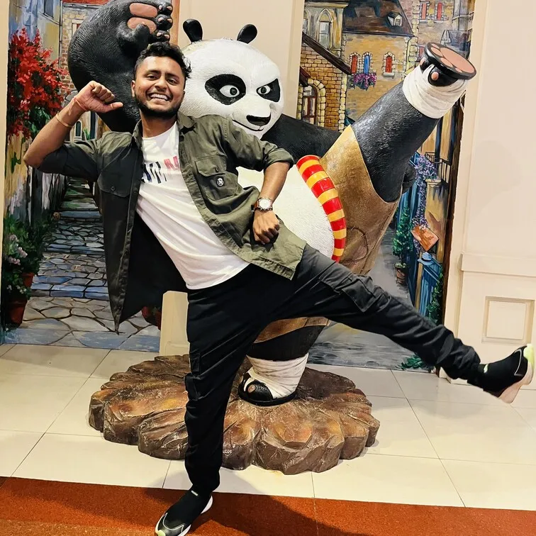 Smiling man posing playfully with one leg raised next to a large Kung Fu Panda statue in an indoor setting.