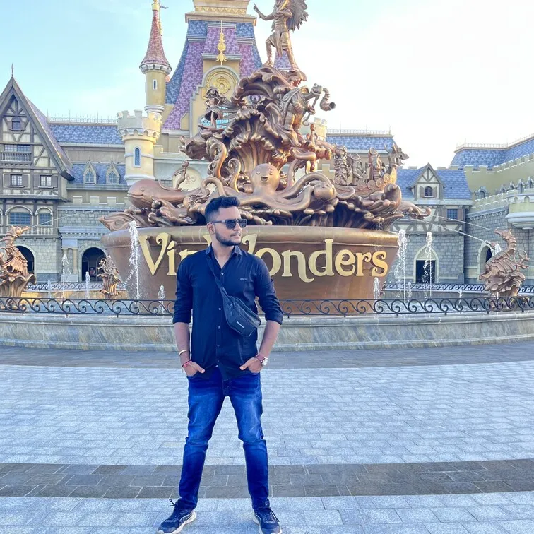 Man wearing sunglasses and dark clothes standing in front of a ornate fountain with 'Vism Wonders' signage and castle-like buildings.