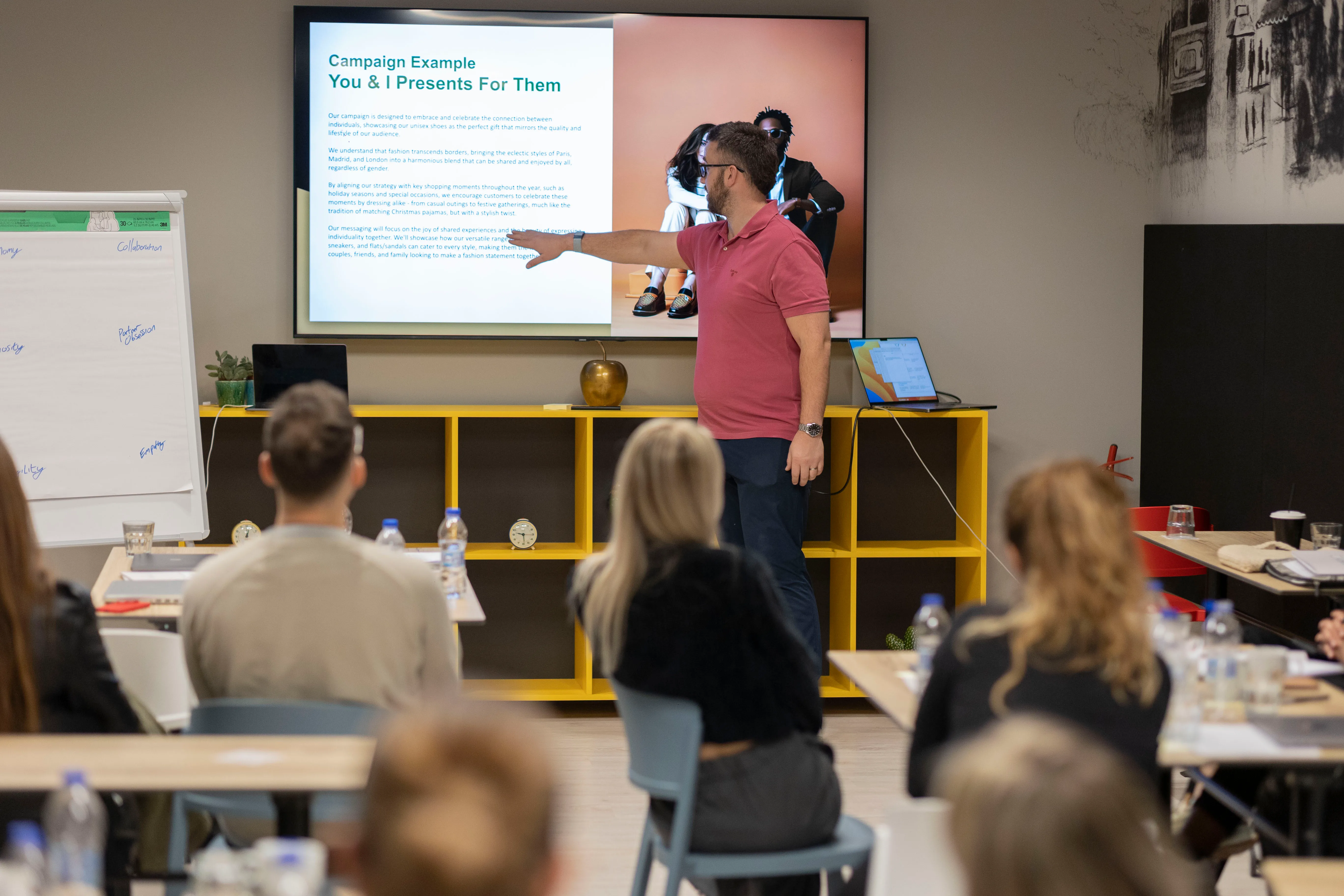 Man in a pink shirt wearing a face mask giving a presentation to an audience in a classroom, pointing at a screen displaying a campaign example titled 'You & I Presents For Them'.