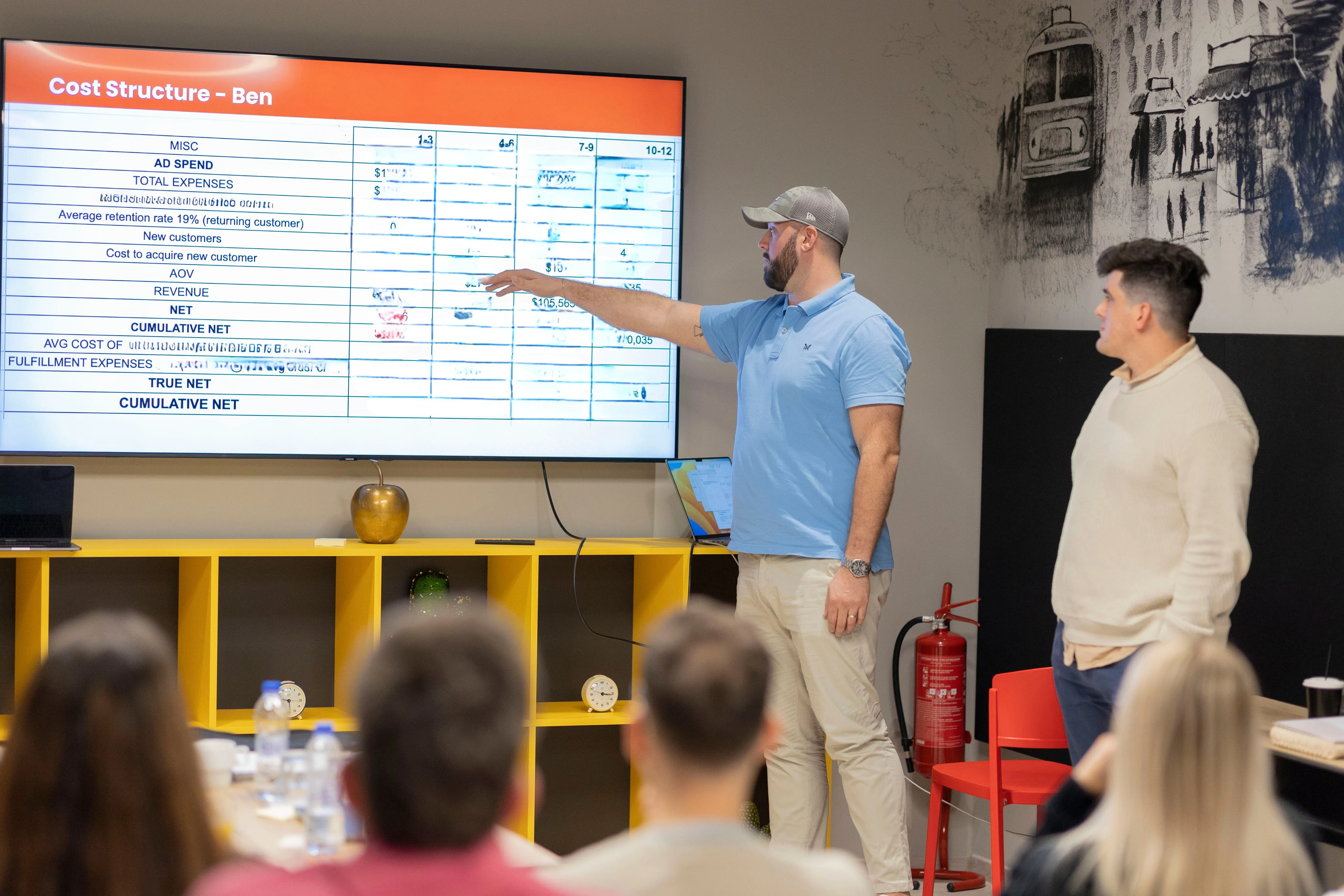 Man in a blue polo and cap pointing at a large screen displaying a cost structure table during a presentation with an audience watching.