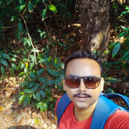 Man wearing sunglasses and a red shirt with a blue backpack standing outdoors in front of lush green foliage and a tree trunk.