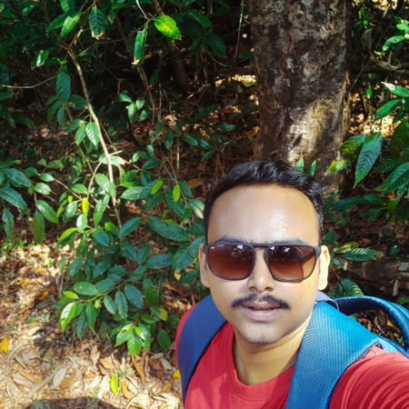 Man wearing sunglasses and a red shirt with a blue backpack standing outdoors in front of lush green foliage and a tree trunk.