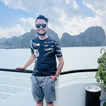 Smiling man wearing sunglasses, black printed t-shirt, and gray shorts standing on a boat deck with mountainous islands and cloudy sky in the background.