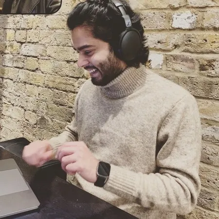 Smiling man wearing a beige turtleneck sweater and headphones, sitting at a table working on a laptop.