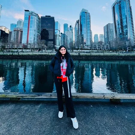 Woman in black jacket and white sneakers standing on a waterfront promenade with tall modern buildings reflected in the water behind her.