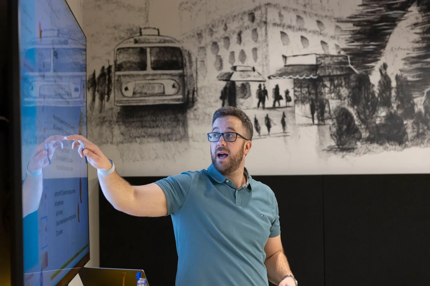 Man in teal polo shirt and glasses pointing at text on a large screen during a presentation in a room with black and white mural background.