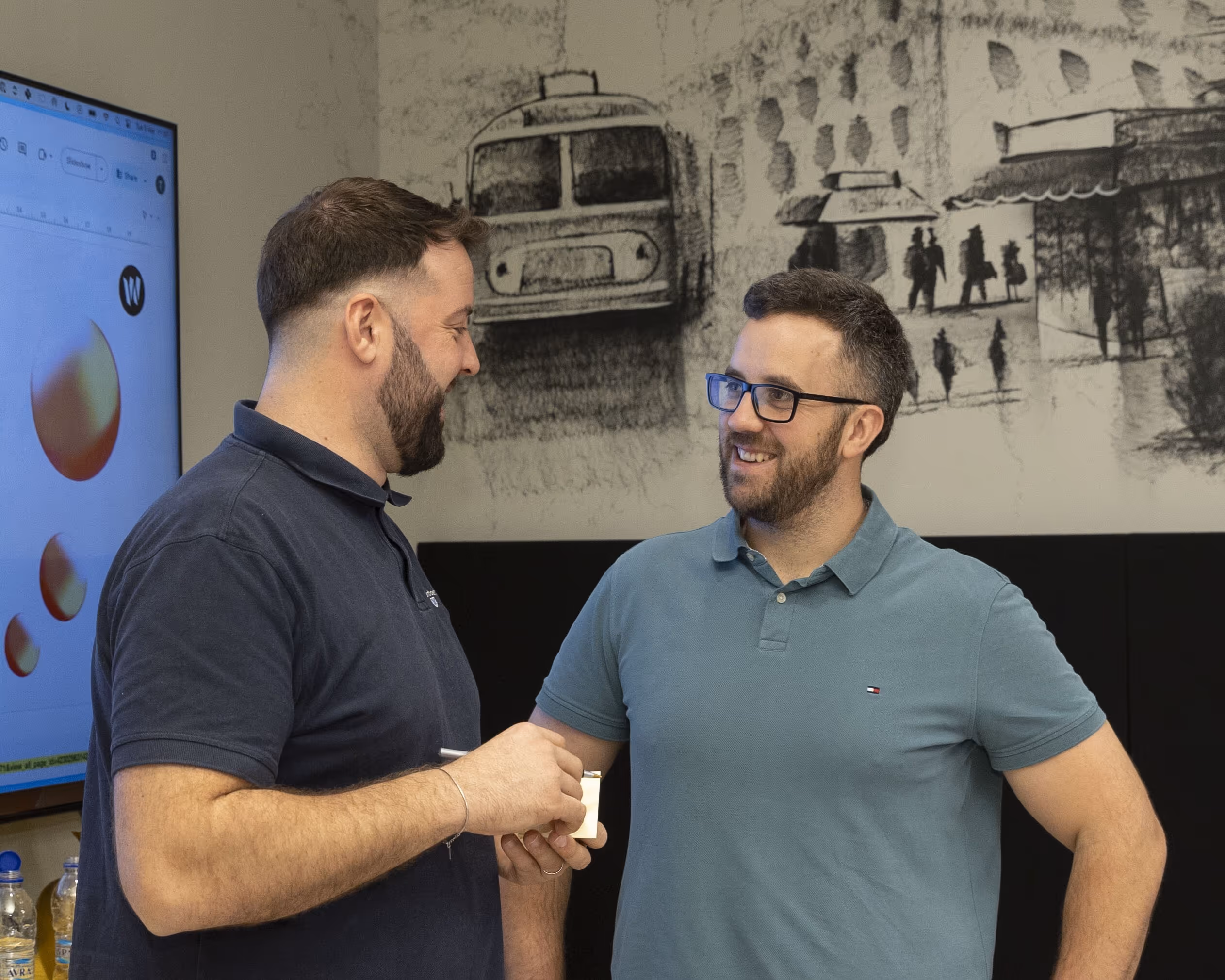 Two men smiling and having a conversation in an office, with a screen displaying 3D shapes and a wall mural in the background.