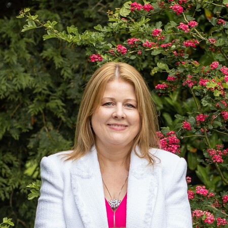 Smiling woman with blond hair wearing a white jacket and pink top, standing outdoors near green foliage and pink flowers.