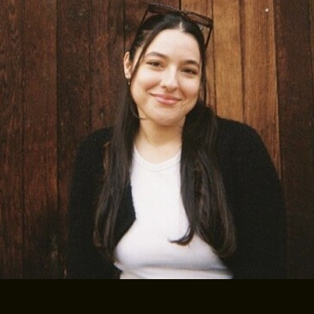 Smiling woman with long dark hair wearing a white top and black cardigan, standing in front of a wooden wall.