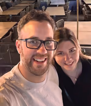 Smiling man with glasses and woman taking a selfie in a restaurant with tables and chairs in the background.